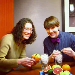 2 instructional designers surrounded by fruit, putting citrus twists into glasses of ice water